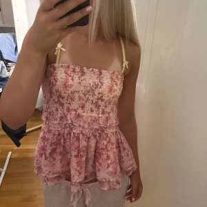 Rosa blommig blus med volang - Supersöt blommig blus i rosa och ljusa toner med smockad överdel och volangig nederdel. Tunna gula axelband med små rosetter ger en gullig touch. Perfekt för varma dagar och somriga vibes. Kan även användas som kjol. 