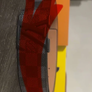 Louis Vuitton rutig svart bälte - Snyggt Louis Vuitton bälte i svart med klassiskt rutigt mönster och stor LV-logga på spännet. Bältet är gjort i skinn och har en stilren design som passar till jeans eller kostymbyxor. Kommer i originalförpackning med blått band.