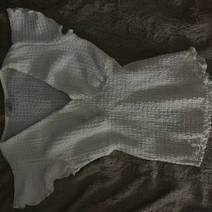 Vit smockad blus med volangärm - Supersöt vit blus från Gina Tricot med smockad struktur och v-ringning. Korta volangärmar och lätt utsvängd nederkant ger en romantisk vibe. Perfekt att styla med jeans eller kjol för en fräsch look.