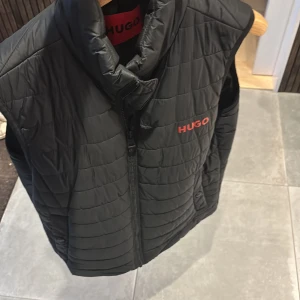 Svart dunväst från Hugo - Snygg svart dunväst från Hugo med röd logga på bröstet. Västen är quiltad med horisontella sömmar, har hög krage och dragkedja framtill. Insidan har en röd detaljficka och tydlig Hugo-etikett. Perfekt för lager-på-lager-stil.