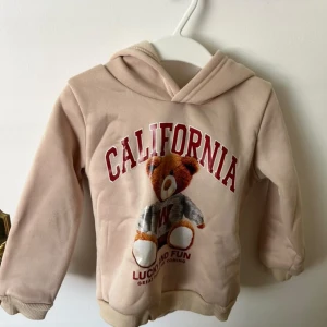 Beige hoodie med björntryck från SHEIN - Söt beige hoodie från SHEIN med huva och tryck av en nallebjörn och texten 'CALIFORNIA' i rött framtill. Tröjan har långa ärmar, ribbade muddar och är gjord i mjuk bomullsblandning. Perfekt för en chill och lekfull look.
