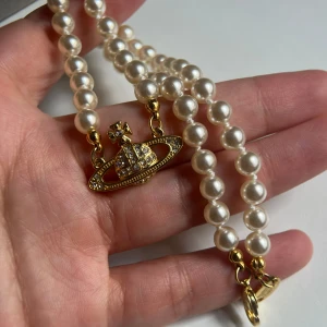 Vivienne Westwood pärlhalsband guld - Ikoniskt pärlhalsband från Vivienne Westwood med vita pärlor och ett guldigt orb-hänge dekorerat med glittrande stenar. Halsbandet har guldfärgade detaljer och klassisk logga på låset. Perfekt statement piece för att lyfta din outfit.