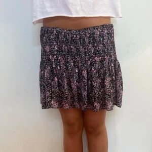 Svart och lila blommig kortkjol - Supersöt kjol med svart botten och lila blommigt mönster. Kjolen har shorts under volangen och är i väldigt bra skick. Kjolen är i stolen L men passar mig som oftast har S-M. Hör av er vid frågor eller funderingar!