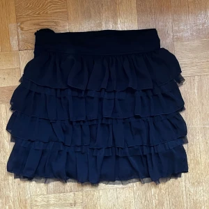 Svart volangkjol med dragkedja - Supersöt svart kortkjol med flera lager med volanger. Kjolen har dragkedja och knapp bak i midjan🩷