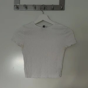 Vit topp - Säljer en vit topp med ”ribbad” struktur och korta ärmar. Toppen har en tight passform och är perfekt för dig som gillar en enkel men trendig stil. Rund halsringning och stretchigt material gör den superbekväm.