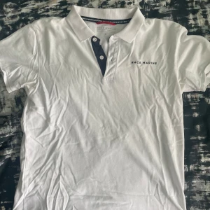 Vit pikétröja från Race Marine - Vit pikétröja från Race Marine med klassisk krage och knäppning framtill. Tröjan har mörkblå detaljer vid knapparna och Race Marine-logga broderad på bröstet. Perfekt för en clean och sportig look.