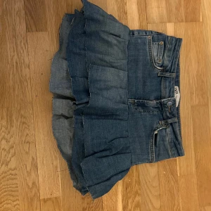 Volangkjol i jeans  - Säljer en blå jeanskjol med volang nertill. Kjolen har klassiska jeansdetaljer som fickor fram och bak, bälteshällor och knappstängning. Den är kort och har en cool, lite ruffad kant längst ner för en avslappnad vibe.