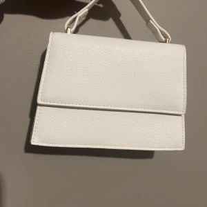 Vit axelväska i skinnlook - Snygg vit axelväska med stilren, fyrkantig form och struktur i materialet. Väskan har ett kort handtag och en avtagbar, justerbar axelrem med guldfärgade detaljer. 