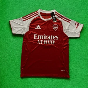 Arsenal Saka 7 matchtröja Adidas - Arsenal hemmatröja med Saka 7-tryck, tillverkad av Adidas i lätt och ventilerande material. Röd bas med vita ärmar och detaljer, klassiska Adidas-ränder på axlarna och klubbmärke på bröstet. Perfekt för dig som vill visa ditt stöd för Arsenal.