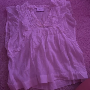 Vit volangblus från Neo Noir - Supersöt vit blus från Neo Noir med volangdetaljer och v-ringning. Blusen är ärmlös och har fina broderade partier samt lätt rynkade partier upptill. Luftigt material som passar perfekt till sommaren och ger en romantisk vibe.