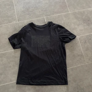 Svart t-shirt från Emporio Armani - Svart t-shirt från Emporio Armani med diskret tryck av loggan upprepat på bröstet. Klassisk passform med rund hals och korta ärmar. Perfekt för en clean och stilren look.