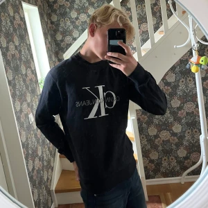 Svart sweatshirt från Calvin Klein Jeans - Svart sweatshirt från Calvin Klein Jeans med stor vit CK-logga framtill. Klassisk rund halsringning och långa ärmar. Tröjan är i mjuk bomullsmix och har en relaxed passform som är perfekt till jeans eller joggers.