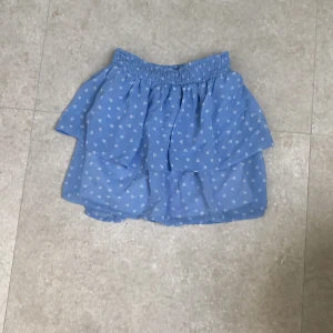 Blå blommig volangkjol Kappahl - Supersöt blå kortkjol från Kappahl med små vita blommor och volang. Kjolen har resår i midjan och är gjord i ett lätt och luftigt material, perfekt för varma dagar. Passar dig som gillar en feminin och trendig stil.