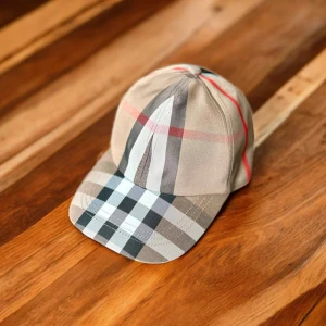 Burberry rutig keps i bomull - Snygg keps från Burberry i klassiskt rutigt mönster med beige, svart, vitt och röda detaljer. Justerbar rem baktill med metallspänne där Burberry-loggan är ingraverad. Tillverkad i bomull och perfekt för dig som vill ha en stilren accessoar med lyxig känsla.