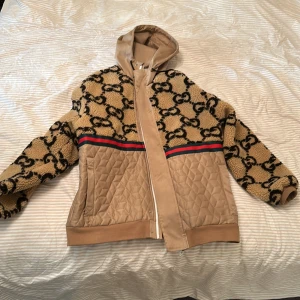 Beige teddyjacka från Gucci med huva - Säljer en beige teddyjacka från Gucci med ikoniskt GG-mönster i svart, quiltade detaljer och klassiska röd-gröna ränder. Jackan har huva, dragkedja och är fodrad med mesh på insidan. Perfekt för dig som vill sticka ut med lyxig streetstyle.