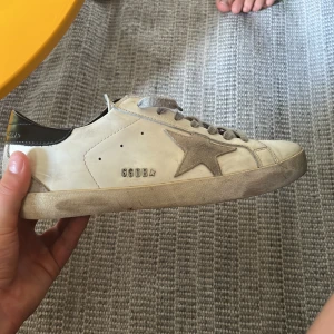 Golden Goose Superstar sneakers vit/grå - Golden Goose Superstar sneakers i vitt skinn med grå mockadetaljer och ikonisk stjärna på sidan. Grå snörning, svart häl och platt sula. Snygg, avslappnad look med coola vintage-vibbar. Perfekta för dig som gillar streetstyle och italiensk design.