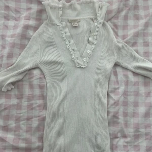 Vit ribbad stickad tröja H&M - Supersöt vit stickad tröja från H&M med ribbad struktur, v-ringning med volangkant och knappar framtill. Tröjan har långa ärmar och en feminin, figurnära passform. Perfekt för dig som gillar romantiska detaljer och vill ha en stilren look.