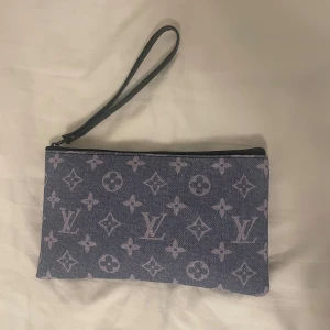  blå kuvertväska  - Snygg blå kuvertväska med klassiskt monogrammönster i vitt. Väskan har en handledsrem i svart skinn och stängs med dragkedja. Perfekt storlek för det viktigaste och enkel att bära med sig. Aldrig använd är 21x14 cm.