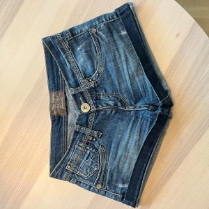Jeansshorts från Stradivarius - Snygga blå jeansshorts från Stradivarius med coola detaljer som kontrastsömmar och dekorativa nitar. Bakfickorna har broderade mönster och en patch i ormskinnsimitation. Klassisk femficksmodell och låg midja. Perfekta för sommaren! Jag kan gå ner i pris vid snabb affär!! Populära så var snabba!