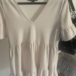 Ljus volangklänning från Rebellious - Supersöt ljus kortklänning från Rebellious med v-ringning, volangdetaljer och korta ärmar. Klänningen har en flowy passform och är tillverkad i ett mjukt, lätt material som känns skönt mot huden. Perfekt för dig som gillar en romantisk och trendig look.