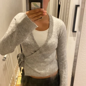 Grå omlottkofta från Gina Tricot - Mysig grå omlottkofta från Gina Tricot i ullblandning. Koftan har lång ärm, knyt i sidan och en mjuk, fluffig känsla. Bra skick. Använd mycket lite. 