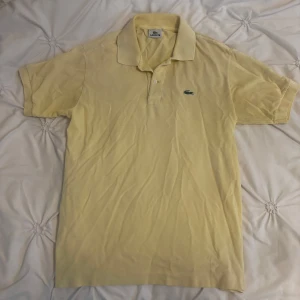 Lacoste T-shirt - Klassisk gul pikétröja från Lacoste med broderad grön krokodil på bröstet. Tröjan har korta ärmar, krage och knäppning med två knappar. Tillverkad i bomull för en skön och luftig känsla. Perfekt för dig som gillar stilren och sportig look.