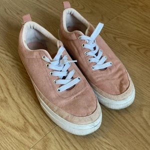 Rosa mockasneakers med vit sula - Snygga sneakers i ljusrosa mocka med vita snören och vit platt sula. Skorna har rund tå och diskreta detaljer i tyg runt hälen. Perfekta för dig som gillar en stilren och fräsch look med en touch av färg.
