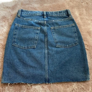 Blå jeanskjol från H&M - Snygg blå jeanskjol från H&M i klassisk femficksmodell med rå kant nertill. Kjolen har bälteshällor, knapp och dragkedja framtill samt två stora bakfickor. Perfekt för dig som gillar en enkel och tidlös denimlook.