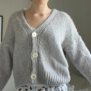 Ljusgrå stickad kofta med stora knappar i mohair - Supermysig ljusgrå stickad kofta med oversized passform och stora vita knappar framtill. Djup v-ringning och ribbade muddar. Tillverkad i en mjuk mix av mohair, ull, nylon och akryl. Perfekt att slänga på sig när det är kyligt ute. Knapparna är fastsydda själv men de går bara att klippa av och byta ut ifall man inte gillar hur de ser ut eller om de går av.💗