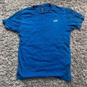 Blå t-shirt från Vans - En klassisk blå t-shirt från Vans med liten vit logga på bröstet. T-shirten har rund halsringning och korta ärmar. Tillverkad i 100% bomull för en skön och avslappnad känsla. Perfekt till jeans eller shorts för en enkel streetstyle.