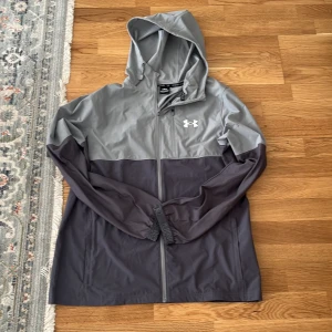 Grå vindjacka från Under Armour - Snygg tvåfärgad vindjacka från Under Armour i grått och mörkgrått. Jackan har huva, hel dragkedja och elastiska muddar. Perfekt för blåsiga dagar och sportiga outfits. Stort Under Armour-logga på bröstet för extra stilpoäng.