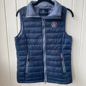 Blå dunväst från Mountain Horse - Snygg blå dunväst från Mountain Horse med quiltad design och dragkedja framtill. Västen har två fickor med dragkedja och en broderad logga på bröstet. Västen har även två innerfickor. Har använts endel och finns tecken på slitage men fortfarande fin, men ej nyskick.Perfekt för lager-på-lager och passar till många outfits.
