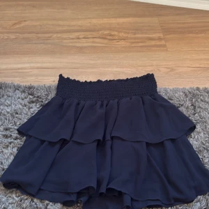 Mörkblå volangkjol med resårmidja - Supersöt mörkblå kortkjol med bred smockresår i midjan och flera lager volanger. Kjolen har en flowy känsla och passar perfekt till en trendig look. Materialet känns lätt och luftigt, perfekt för varmare dagar.