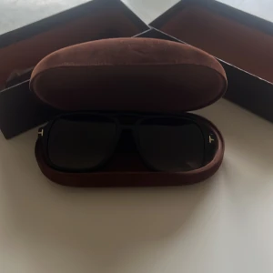 Svarta solglasögon från Tom Ford - Snygga svarta solglasögon från Tom Ford med klassisk fyrkantig form och diskreta gulddetaljer vid skalmarna. Kommer i original brun sammetsask och box. Perfekta för dig som vill ha en stilren och lyxig look.