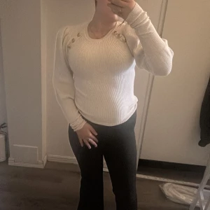 Vit ribbad stickad tröja med puffärm - Supersnygg vit stickad tröja med ribbad struktur och puffärmar. Tröjan har dekorativa guldknappar vid axlarna som ger en extra touch. Långärmad och figurnära passform, perfekt till både jeans och kjol. Stilren och trendig för dig som gillar detaljer.