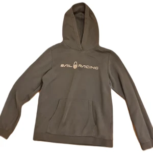 Grå hoodie från Sail Racing - Snygg grå hoodie från Sail Racing med klassisk logga framtill. Tröjan har huva, magficka och långa ärmar. Perfekt för dig som gillar en enkel och stilren look. Materialet är mjukt och skönt, troligen bomullsmix.