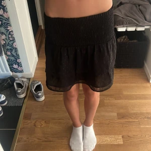 Svart kortkjol med volang - Supersöt svart kortkjol med bred smock i midjan och volangig nederdel. Kjolen har små diskreta prickar i tyget .  Sitter skönt och är lätt att matcha med olika toppar.