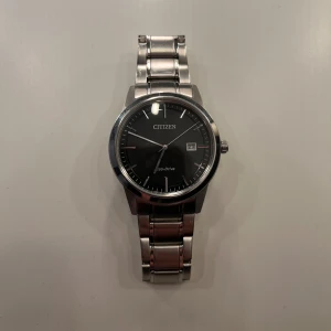 Citizen Eco-Drive silverklocka - Stilren Citizen Eco-Drive klocka med svart urtavla och diskreta linjer. Klockan har datumvisning och ett klassiskt armband i silverfärgad metall. Perfekt för dig som gillar en clean och tidlös look med modern teknik.