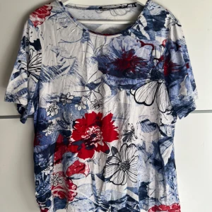 Blommig t-shirt från Zhenzi - Snygg t-shirt från Zhenzi med stort blommönster i rött, blått och svart på vit botten. T-shirten har korta ärmar, rund halsringning och är gjord i mjuk bomull. Detalj med små silvriga nitar vid halsen. Perfekt för dig som gillar färg och mönster!