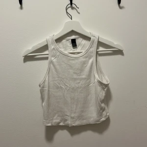 Vit ribbad croppad linnetopp - Säljer en enkel och fräsch vit croppad linnetopp. Toppen är ribbad och ärmlös med rund halsringning, perfekt för varma dagar och passar dig som gillar en clean och stilren look. Den är något nopprig på baksidan. 
