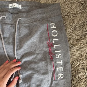 Grå mjukisbyxor Hollister London S - Grå mjukisbyxor från Hollister med broderad text 'Hollister London' längs benet och en liten brittisk flagga vid midjan. Byxorna har dragsko i midjan och är gjorda i mjukt bomullsmaterial. Perfekta för chill dagar eller när du vill vara extra bekväm.