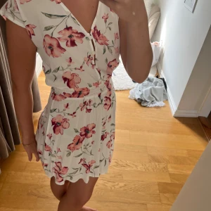 Vit blommig kortklänning med v-ringning - Superfin vit kortklänning med rosa och röda blommor, v-ringning och knappar framtill. Klänningen har resår i midjan och korta ärmar, perfekt för varma dagar. Lätt och luftig, passar dig som gillar romantisk stil.