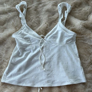 Vit linnetopp med volangband och snörning - Supersöt vit linnetopp med broderade hål och volangiga axelband. Toppen har v-ringning och snörning både fram och bak för en extra snygg detalj. Perfekt för varma dagar och ger en somrig vibe. 