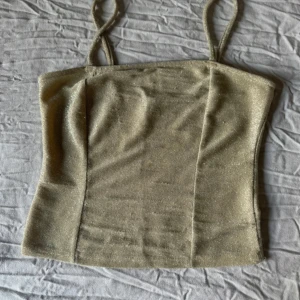 Glittrig guldig linne från Gina Tricot - Supersnygg glittrig guldig linne från Gina Tricot med smala axelband och figurnära passform. Perfekt att styla med jeans eller kjol för en trendig look. Materialet är stretchigt och skimrar snyggt i ljuset. Har blivit lite nopprig. Men inget som syns ifall man inte kollar ordentligt.