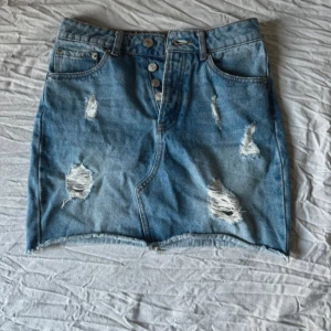 Blå jeanskjol med slitningar från BikBok - Snygg blå jeanskjol från BikBok i kort modell med slitna detaljer och rå kant nertill. Kjolen har knappar framtill, klassiska fickor och bälteshällor. Perfekt för en chill och trendig look.