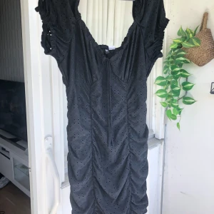 Svart kort klänning med broderi - Supersöt svart kortklänning med broderade detaljer och små hålmönster. Klänningen har rynkade partier framtill, korta puffärmar som man kan sätta ”vanligt” och som off shoulder, och en söt knytning vid halsen. 