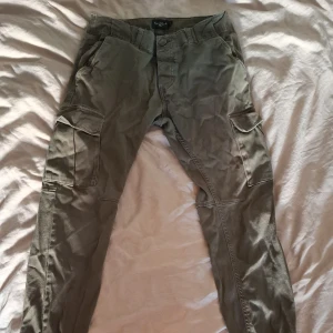 Olivgröna cargopants från Saligia XS - Säljer ett par olivgröna cargopants från Saligia i storlek XS. Byxorna har flera stora fickor på sidorna och bak, klassisk cargomodell och är tillverkade i bomull. Perfekta för en avslappnad och cool streetstil.