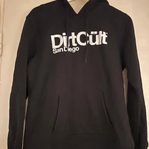 Svart DirtCult hoodie med tryck - Svart hoodie från DirtCult med stort vitt tryck på bröstet där det står 'DirtCult San Diego'. Klassisk modell med huva och känguruficka framtill. Perfekt för dig som gillar streetwear och vill ha en chill look. Storlek XL men är som en M