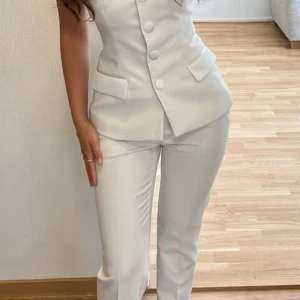 Vit figursydd väst med stora knappar - Snygg vit väst med figurnära passform och stora klädda knappar framtill. Västen är ärmlös, har en hjärtformad urringning och två dekorativa fickor. Materialet är slätt och ger en elegant känsla. Perfekt för att lyfta en outfit med en modern touch.