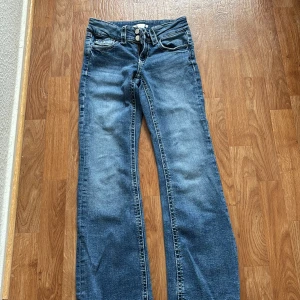 Blå bootcut jeans från Gina Tricot - Snygga blå jeans från Gina Tricot i klassisk bootcut-modell. Jeansen har normal passform, fem fickor och dubbla knappar i midjan. Tillverkade i jeansmaterial med ljusare tvätt och tydliga sömmar. Perfekta för dig som gillar en avslappnad och trendig look.